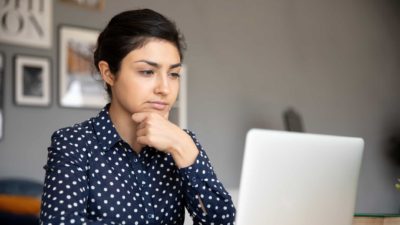 A young woman sits at her desk in deep contemplation with her hand to her chin while seriously considering information she is reading on her laptop.